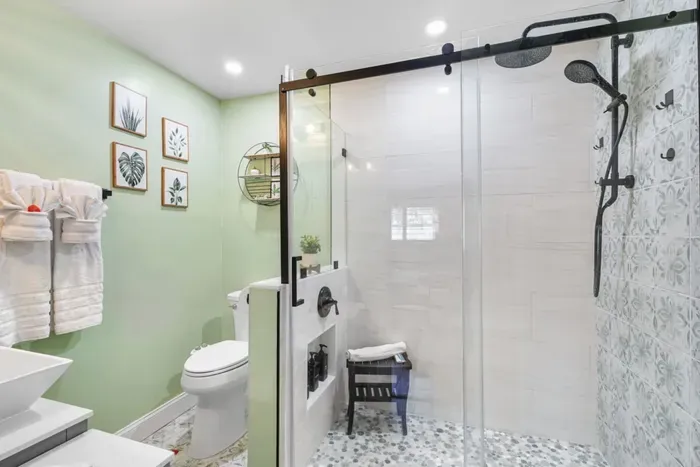 The spa-inspired ensuite bathroom in Palm Paradise features a walk-in glass rain shower with dual shower heads, pebble tile flooring, botanical décor, and a sliding barn-style glass door.