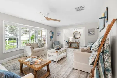 The spacious lounge area at Twin Oaks Coastal Luxury, featuring neutral coastal furnishings, a round woven mirror, pelican and heron artwork, and large plantation-shuttered windows filling the room with natural Florida light.