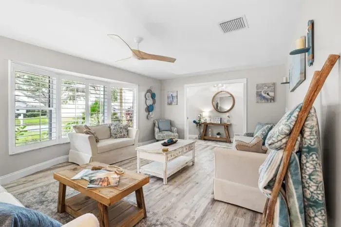 The spacious lounge area at Twin Oaks Coastal Luxury, featuring neutral coastal furnishings, a round woven mirror, pelican and heron artwork, and large plantation-shuttered windows filling the room with natural Florida light.