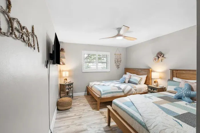 Another view of Castaway's Respite at Twin Oaks — two queen beds with coastal quilts, warm bedside lighting, a smart TV, and shell wind chime accents that invite you to relax and unwind.