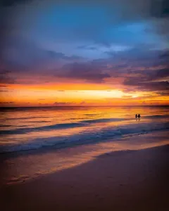 A breathtaking Gulf Coast sunset just minutes from Twin Oaks Coastal Luxury — vivid orange and violet skies reflected over the water's edge, with two silhouettes wading in the warm Gulf waves.