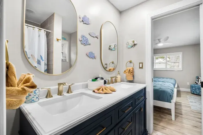 A beautifully updated Jack-and-Jill style bathroom at Twin Oaks Coastal Luxury, featuring a navy double vanity with gold hardware, dual oval mirrors, and playful coastal fish wall art — accessed from Sailor's Cabin.