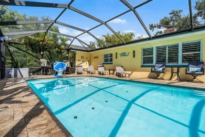 A sparkling view of the full pool at Twin Oaks Coastal Luxury, with travertine decking, a row of sun loungers along the yellow stucco house wall, a pool basketball hoop, and lush majestic oaks towering above the screened enclosure.