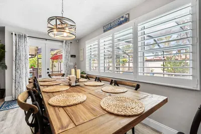 The sunlit dining room at Twin Oaks Coastal Luxury seats the full group around a rustic wood farmhouse table, with an 'Indian Shores' sign above the plantation-shuttered windows and French doors opening to the pool patio.
