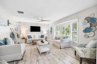 A bright, airy second living space with white sofas, a distressed wood coffee table, starfish accents, and large plantation shuttered windows overlooking lush Florida greenery.