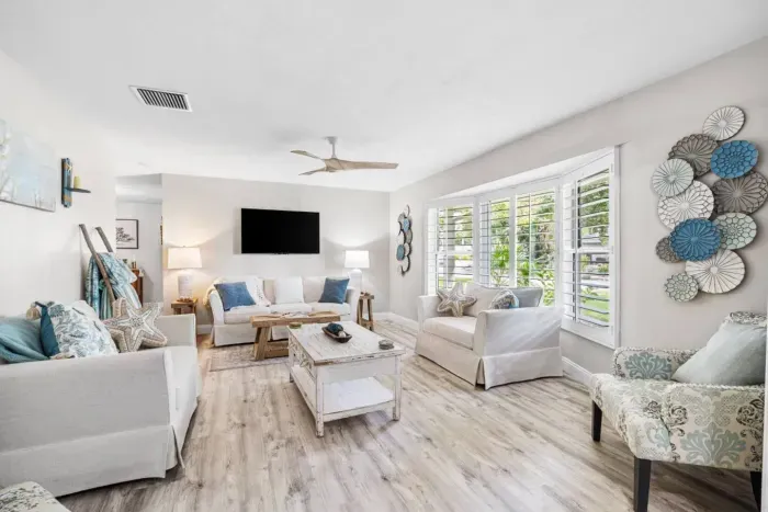 A bright, airy second living space with white sofas, a distressed wood coffee table, starfish accents, and large plantation shuttered windows overlooking lush Florida greenery.