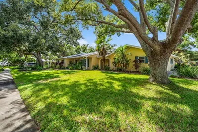 The sunny exterior of Twin Oaks Coastal Luxury — a charming Florida home with yellow stucco, classic brick accents, and lush tropical landscaping including towering palms that set the island vibe before you even step inside.
