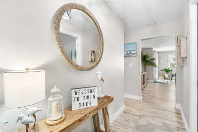 The welcoming entryway at Twin Oaks Coastal Luxury, featuring a 'Welcome to Twin Oaks' light-box sign, a driftwood console table, woven round mirror, and shorebird décor — setting the coastal tone the moment you arrive.