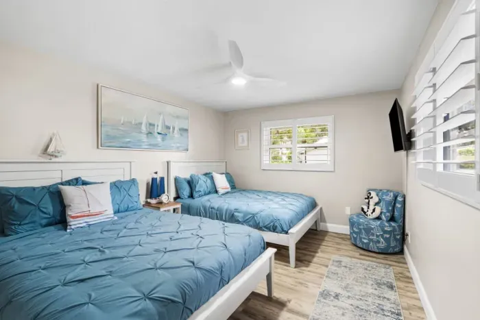 Sailor`s Cabin at Twin Oaks Coastal Luxury: two queen beds dressed in rich blue pinch-pleat bedding, a sailboat painting above the headboard, Smart TV, and a cozy anchor-print accent chair.
