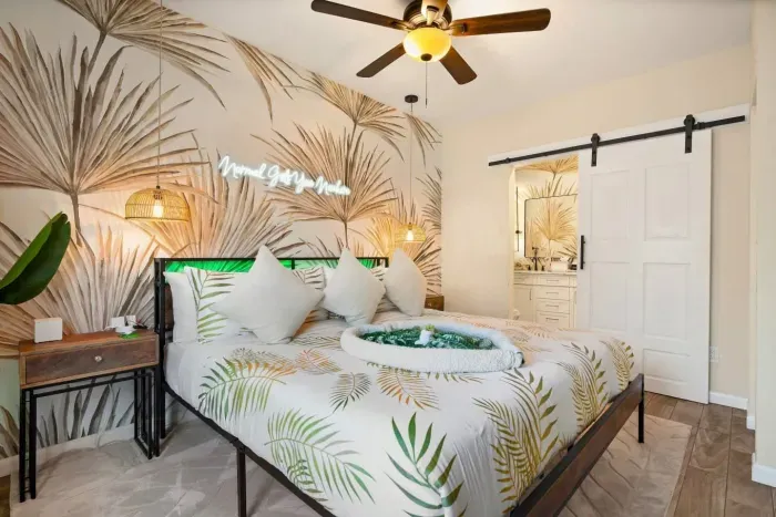 Primary king suite — luxurious coastal bedding, palm-leaf mural, neon accent, woven pendant lights, and barn door entry to private spa ensuite. St. Petersburg, FL vacation rental.