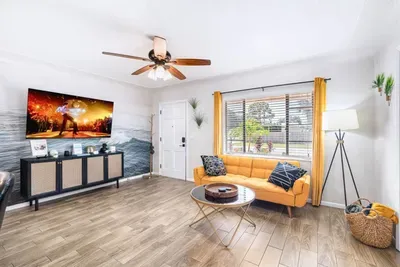 Main living room — 65-inch smart TV, mustard yellow sofa, natural light, and open flow to kitchen. 4-bedroom pool home, St. Petersburg, FL.