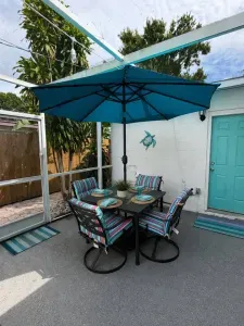 Covered lanai dining area — a four-seat patio table set against the pool enclosure wall, framed by a signature teal door, sea turtle wall art, and tropical plantings.
