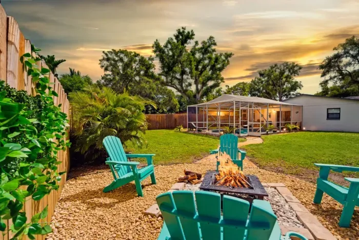 Golden-hour fire pit at Sun Oasis — teal Adirondack chairs, real wood flames, lush tropical palms, and a screened pool glowing in the background. The perfect Florida evening.