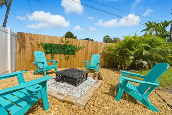Dedicated fire pit area — four teal Adirondack chairs arranged around a wood-burning pit, surrounded by crushed stone, tropical palms, and a green ivy accent wall.