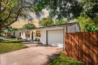 Wide-angle dusk shot of this St. Petersburg vacation rental — white exterior, one-car garage, paver driveway with room for three vehicles, lush mature oak trees, and a welcoming front porch bench.