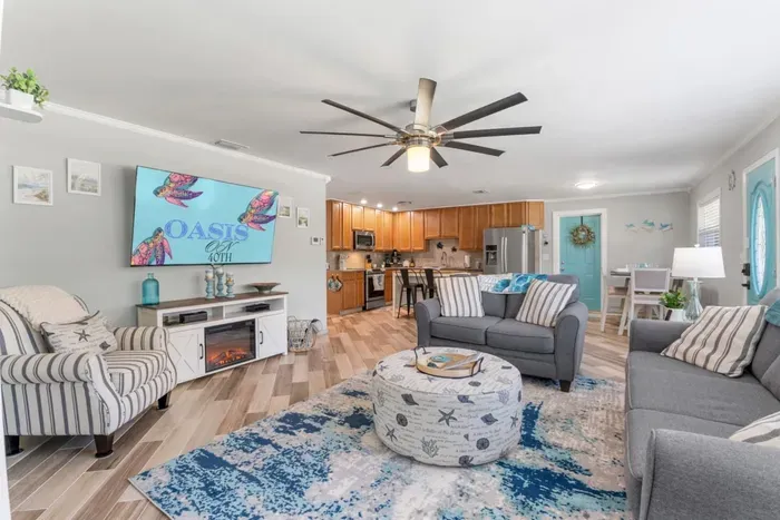 Open-concept living room at Sun Oasis with a large smart TV, fireplace credenza, coastal turtle artwork, beachy blue-and-white decor, and views straight through to the fully equipped kitchen.