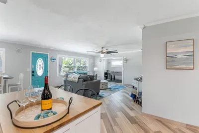 Looking from the butcher-block kitchen island through Sun Oasis's open-plan living and dining rooms — the signature teal front door, coastal art, and the game room entrance all in view.