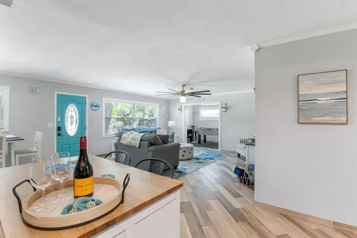 Looking from the butcher-block kitchen island through Sun Oasis's open-plan living and dining rooms — the signature teal front door, coastal art, and the game room entrance all in view.