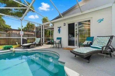 Sun-soaked pool deck at Sun Oasis with multiple loungers, a patio dining set, teal decor, and sliding door access to the primary bedroom — built for all-day relaxing.