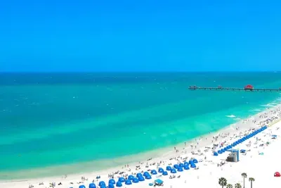 Aerial view of Clearwater Beach, Florida — turquoise Gulf water, white sand, blue beach cabanas, and Pier 60 stretching into the sea. Just 18 miles from this St. Pete vacation rental.