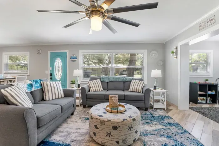 Comfortable coastal living room with two plush sofas, a striped armchair, oversized round ottoman, 65-inch Smart TV, guest book, and easy access to the game room.