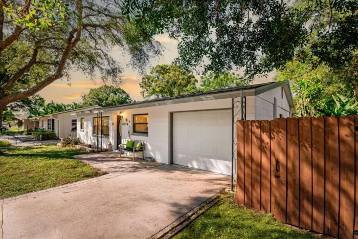 Wide-angle dusk shot of this St. Petersburg vacation rental — white exterior, one-car garage, paver driveway with room for three vehicles, lush mature oak trees, and a welcoming front porch bench.