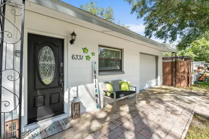 Welcoming front entry of Sun Oasis vacation rental in St. Petersburg, FL — stained glass door, sea turtle wall art, keypad smart lock, paver walkway, and a shaded porch bench set the tone before you even step inside.