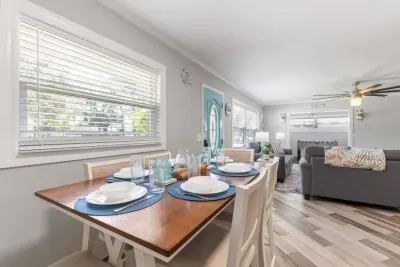 Coastal dining room at Sun Oasis — a farmhouse-style table set for six with blue place settings and teal glassware, teal front door visible beyond, flowing into the living room.