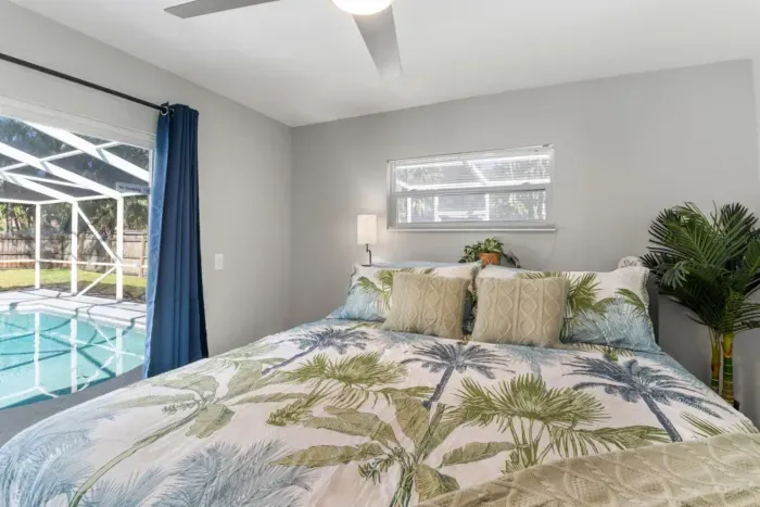 Wake up steps from the pool — the primary bedroom with direct sliding door pool access, coastal luxury bedding, and soft natural light filtering through the screen enclosure.