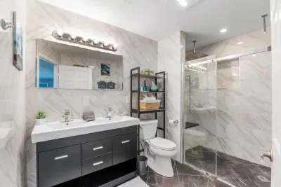Spa-inspired ensuite bathroom — floor-to-ceiling marble tile, oversized double rain shower with frameless glass doors, double vanity, and coastal accent art.