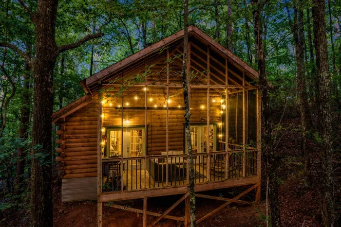 Cozy log cabin in a wooded forest at dusk with warm string lights glowing on a screened-in porch, surrounded by tall trees and lush greenery.