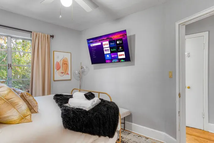 A cozy bedroom with light walls, wood floors, and two large windows. It features a gold-framed bed with white and black bedding, golden pillows, and a wall-mounted TV.