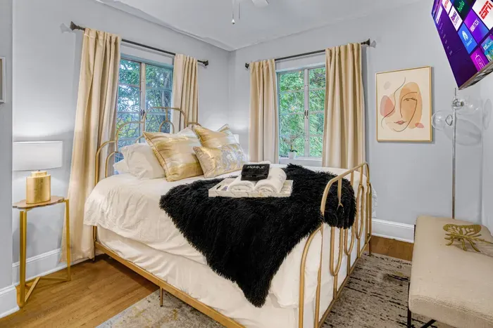 A cozy bedroom with light walls, wood floors, and two large windows. It features a gold-framed bed with white and black bedding, golden pillows, and a wall-mounted TV.