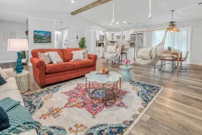A vibrant and open-concept living space featuring a burnt-orange sofa, two cozy hanging hammock chairs, and a stylish rug that ties the room together. The space flows seamlessly into the kitchen and dining area.