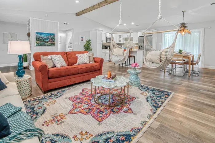 A vibrant and open-concept living space featuring a burnt-orange sofa, two cozy hanging hammock chairs, and a stylish rug that ties the room together. The space flows seamlessly into the kitchen and dining area.