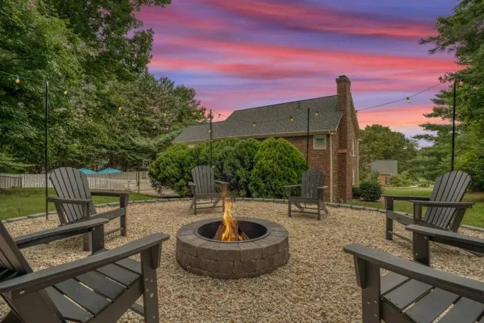 Cozy outdoor fire pit area surrounded by Adirondack chairs, perfect for evening gatherings under string lights and colorful sunset skies.