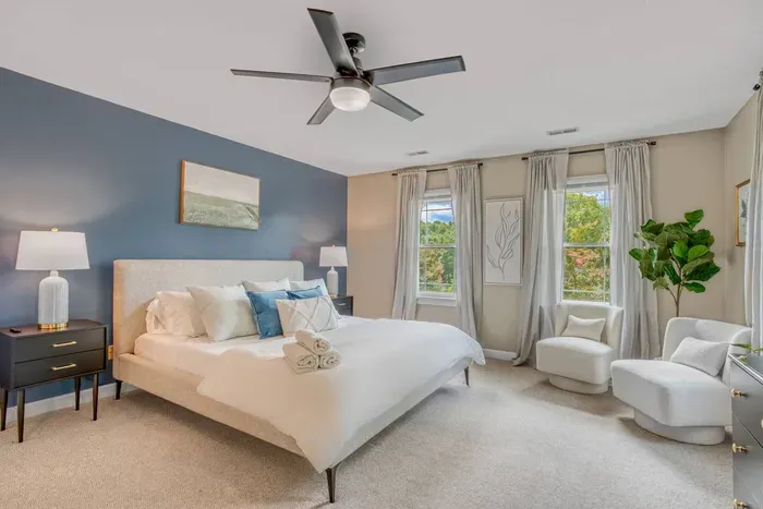 This primary bedroom features a plush king bed, soothing blue accent wall, and large windows that fill the space with natural light and lounge seats.