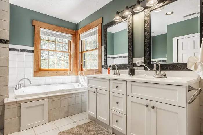A double-sink white vanity with ornate framed mirrors and a deep soaking tub beneath large windows.
