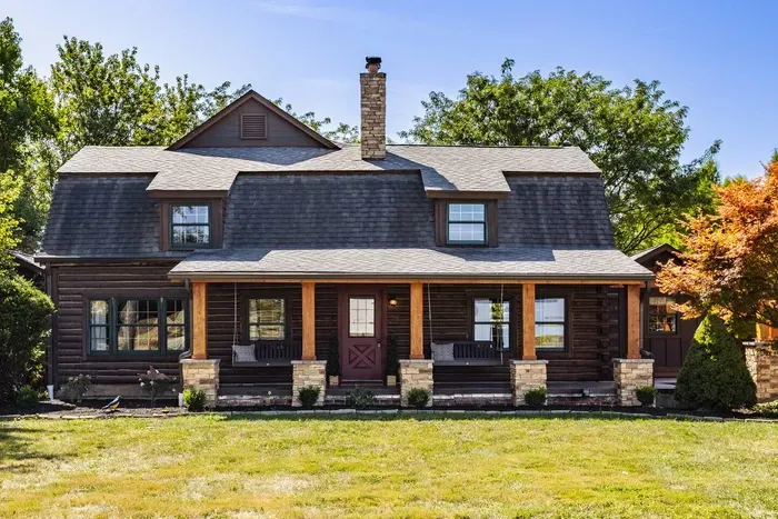 A welcoming front porch with stone pillars, dark wood siding, and a classic pitched roof surrounded by mature trees and a spacious lawn.