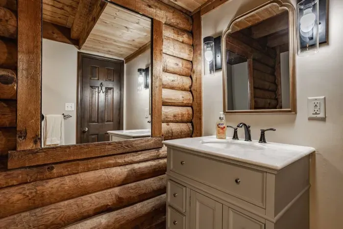 Rustic bathroom with log walls, a gray vanity, white countertop, and an arched mirror accented by modern sconces.