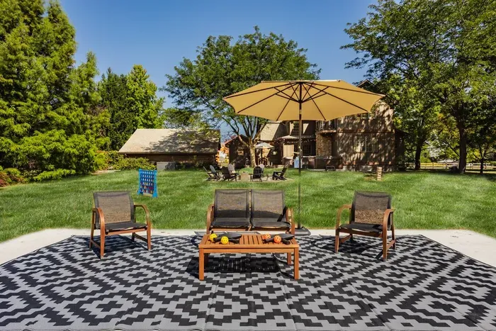 Backyard patio featuring modern outdoor seating under a large tan umbrella, set on a bold geometric rug with a spacious green lawn and trees surrounding a rustic-style home.