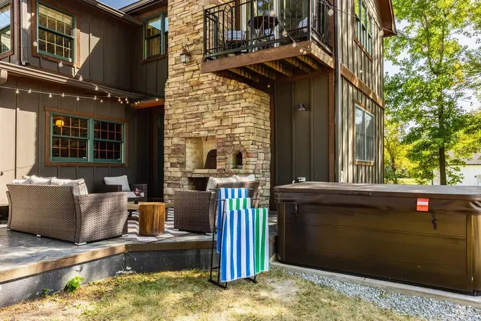 Featuring a sleek hot tub beside the stone fireplace and comfortable wicker seating. The setup is accented by string lights and a balcony overhead.