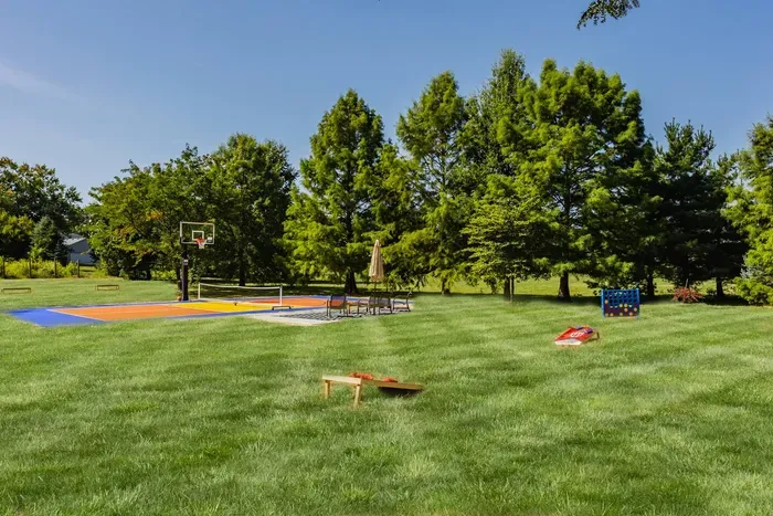 Wide, grassy yard featuring a basketball court, outdoor games, and shaded seating.
