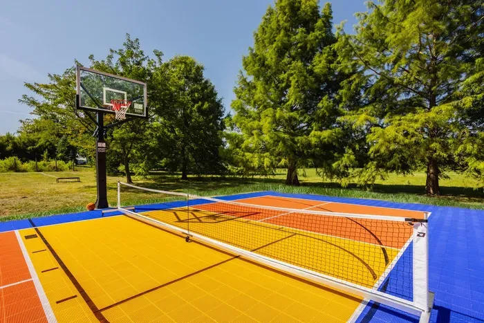 Vibrant multi-sport court featuring a bold blue, orange, and yellow surface, equipped for both basketball and pickleball, surrounded by lush greenery and tall trees.
