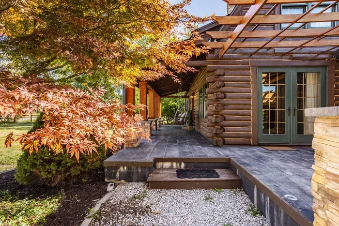 Patio area featuring a log cabin exterior, slate tile flooring, and French doors that open to the outdoors. A pergola and mature landscaping add warmth and charm to this cozy outdoor retreat.