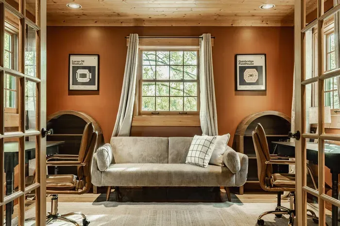 Warm-toned office featuring two wooden desks with chairs, a cozy sofa, and a large window framed by light curtains.
