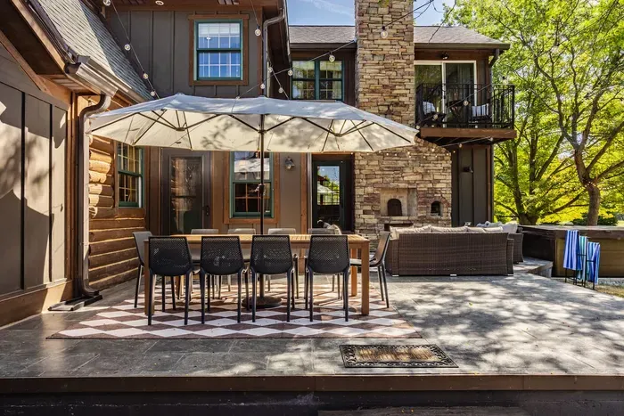 An outdoor patio featuring a long dining table beneath a large umbrella, surrounded by sleek black chairs. String lights add charm overhead, while a cozy seating area and stone chimney complete this inviting space for entertaining or relaxing.