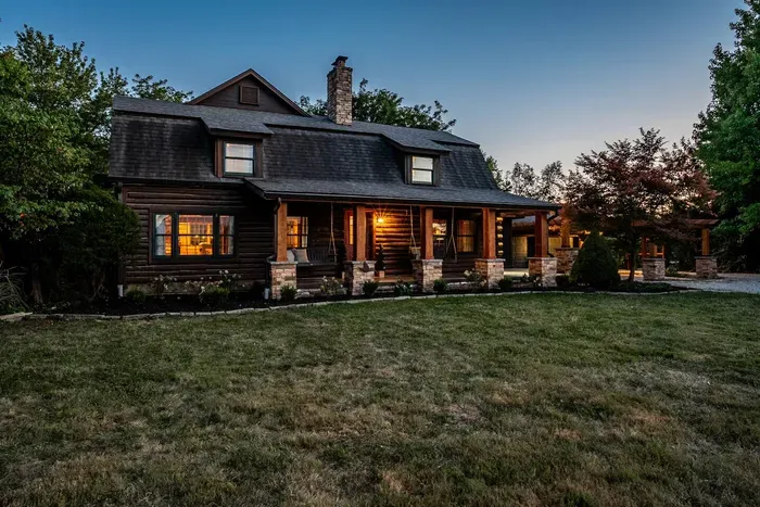 Charming log home with rustic timber architecture, dormer windows, and a spacious covered porch framed by stone columns and surrounded by mature greenery.