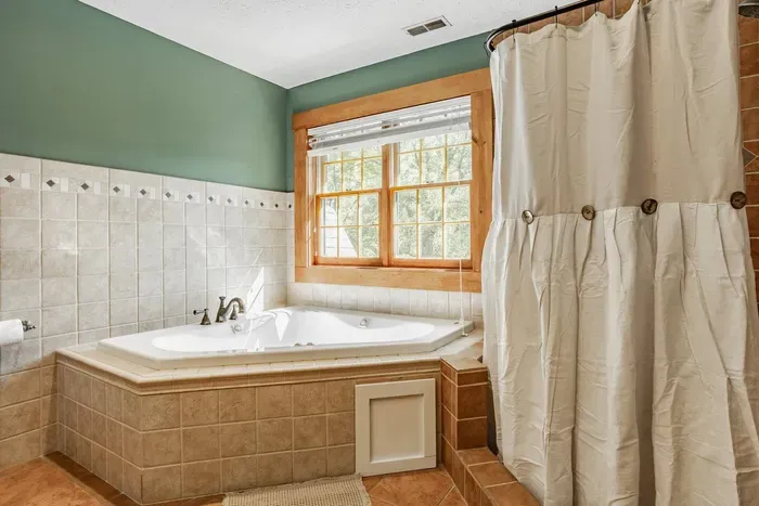 Bright bathroom featuring a spacious corner jetted tub framed by beige tiles and a large wood-trimmed window that lets in plenty of natural light, creating a relaxing and spa-like atmosphere.