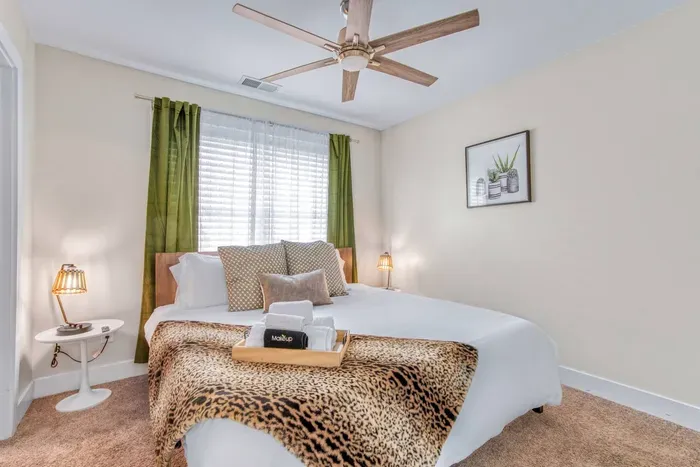 A bright and tranquil bedroom with ceiling fan,  green curtains, soft lighting, and a plush bed accented by a chic leopard print throw.